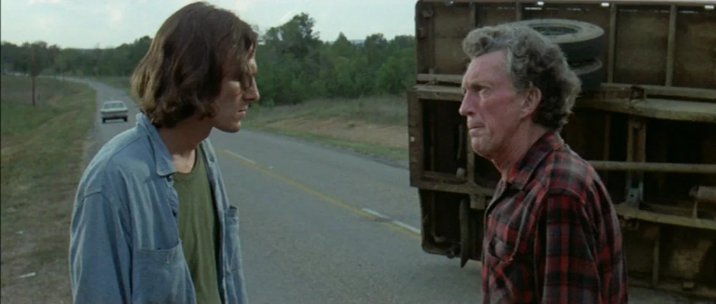 Two Lane Blacktop (1971) movie still: medium — A rural paved road features a large overturned trailer resting on the grassy shoulder. Two men stand…