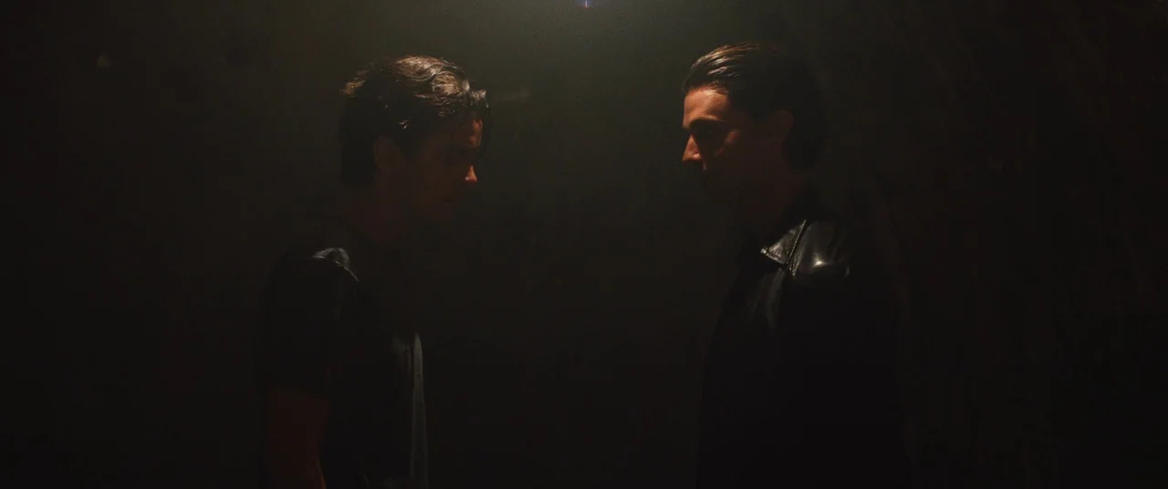 Daniel Isn’t Real movie still: medium — A dimly lit, featureless space obscures the background in heavy shadow. Two young men with dark hair stand…