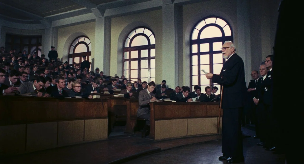 The Damned (1969) movie still: wide — An academic lecture hall features tiered wooden seating filled with students working at desks. A bearded…