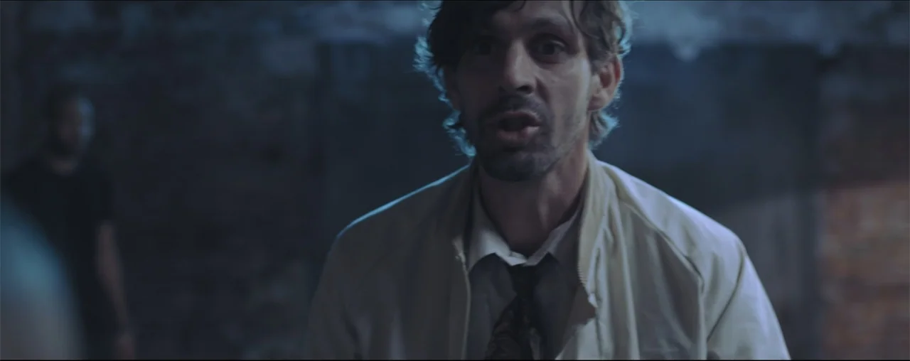 El Último Juego (2025) movie still: medium — A dimly lit, textured space features dark walls and a blurred foreground element. A man in a…