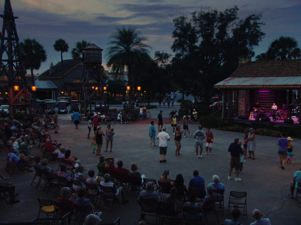 Some Kind Of Heaven (2020) movie still: wide — An open plaza at dusk features a small wooden stage with live performers. Numerous people are seated…