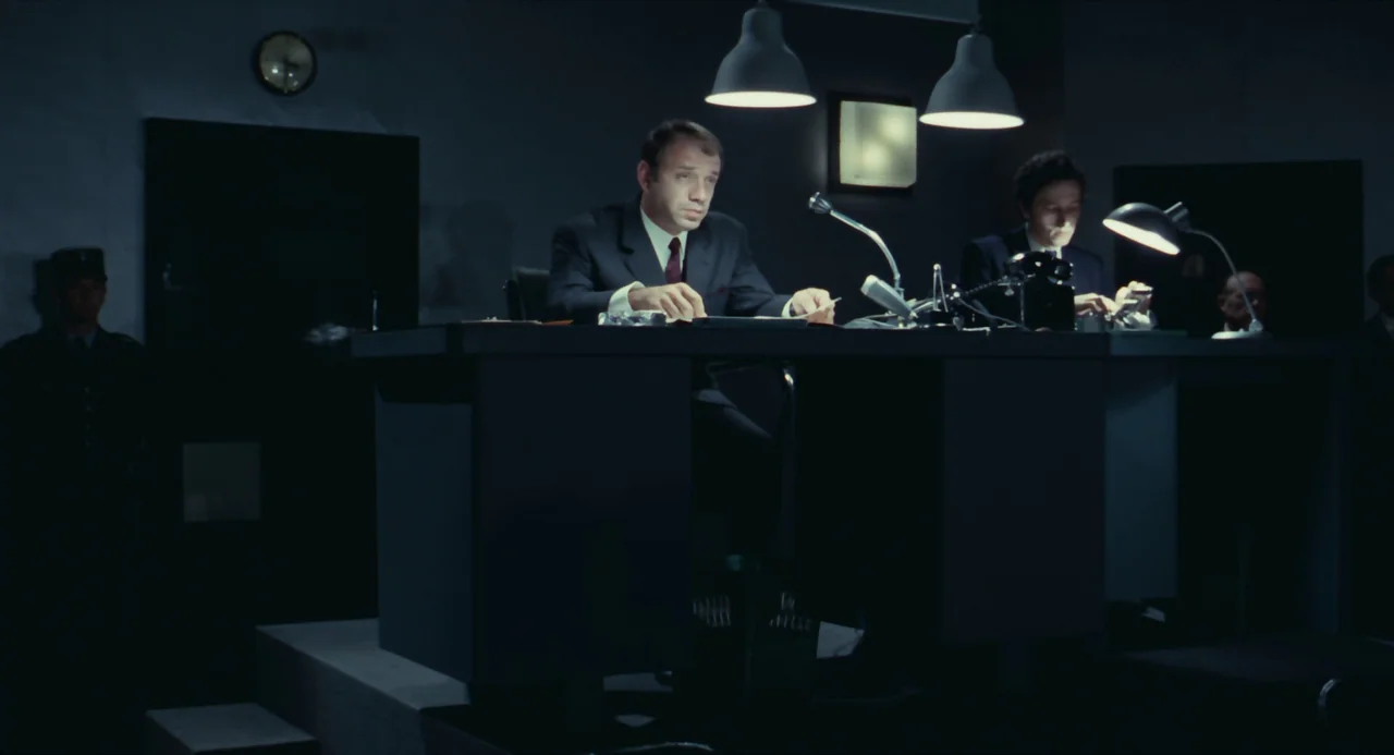 Le Samouraï (1967) movie still: medium — A dimly lit room with multiple desks arranged for official questioning. A man sits at the primary desk…