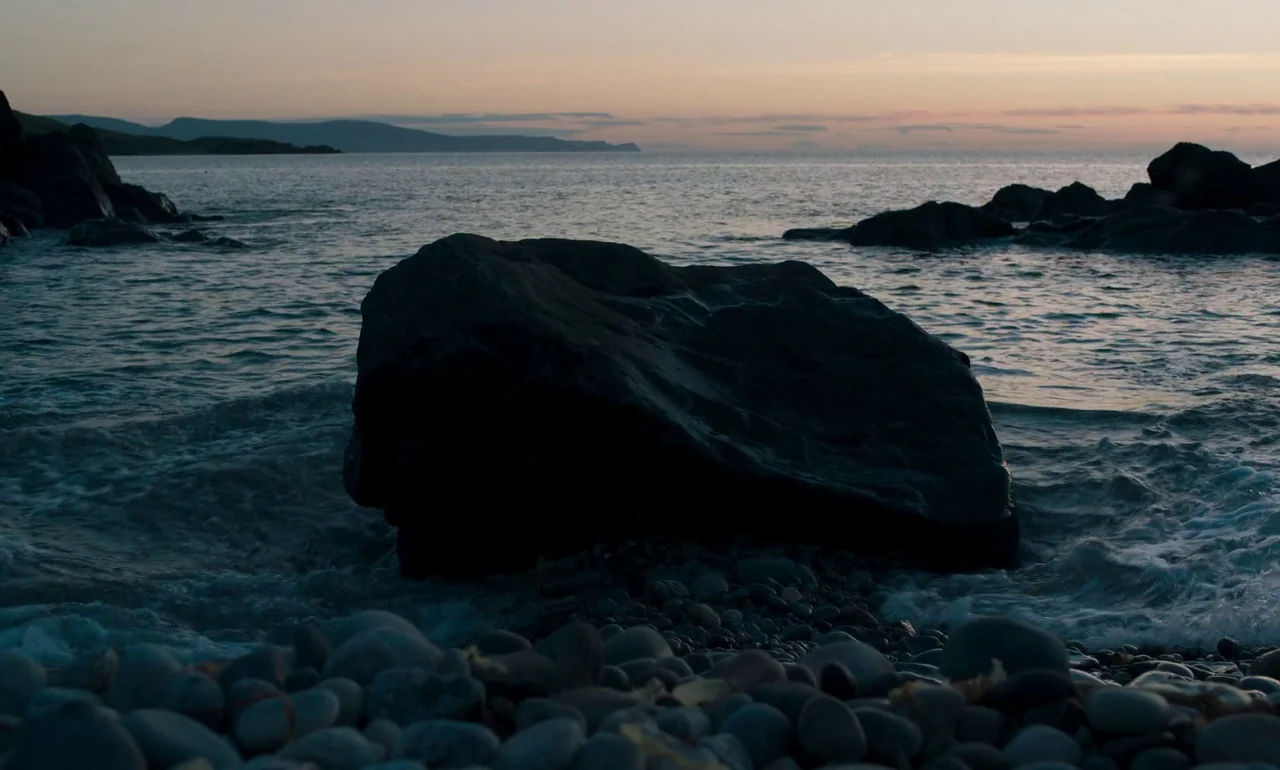 It Is In Us All (2022) movie still: wide — A pebbled beach sits at the edge of a calm, dark ocean at dusk. Large, dark rocks are scattered along the…