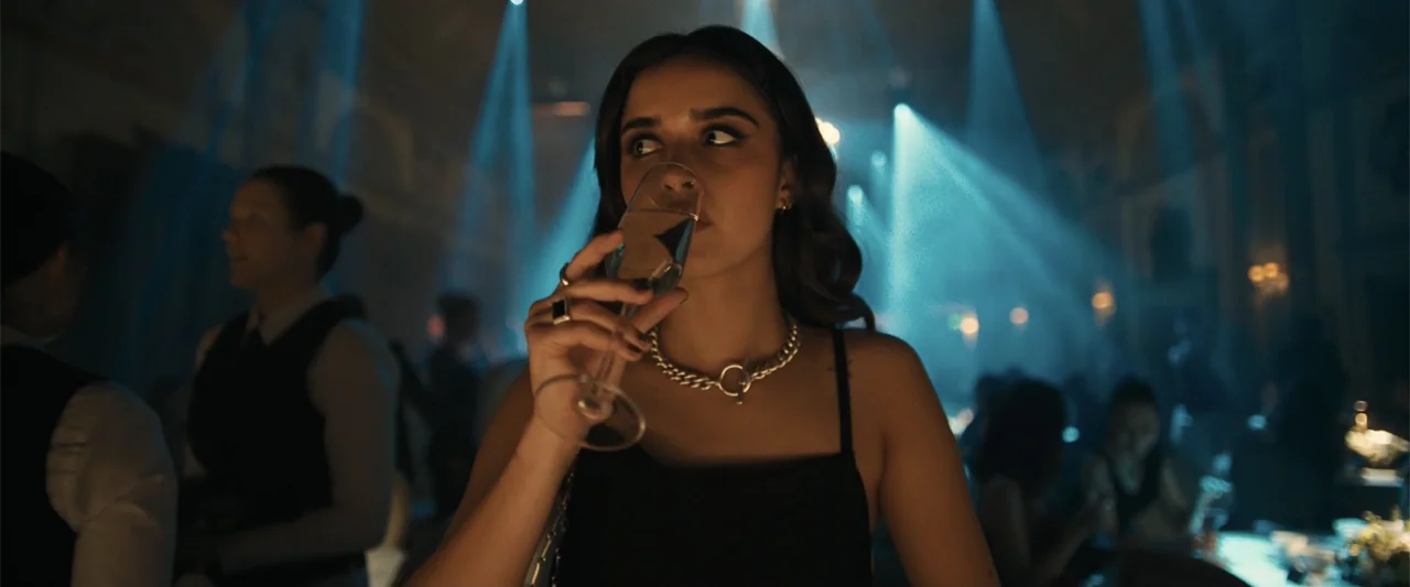 My Fault: London (2025) movie still: medium — A dimly lit club interior features blue spotlights cutting through ambient haze. A woman drinks from a…
