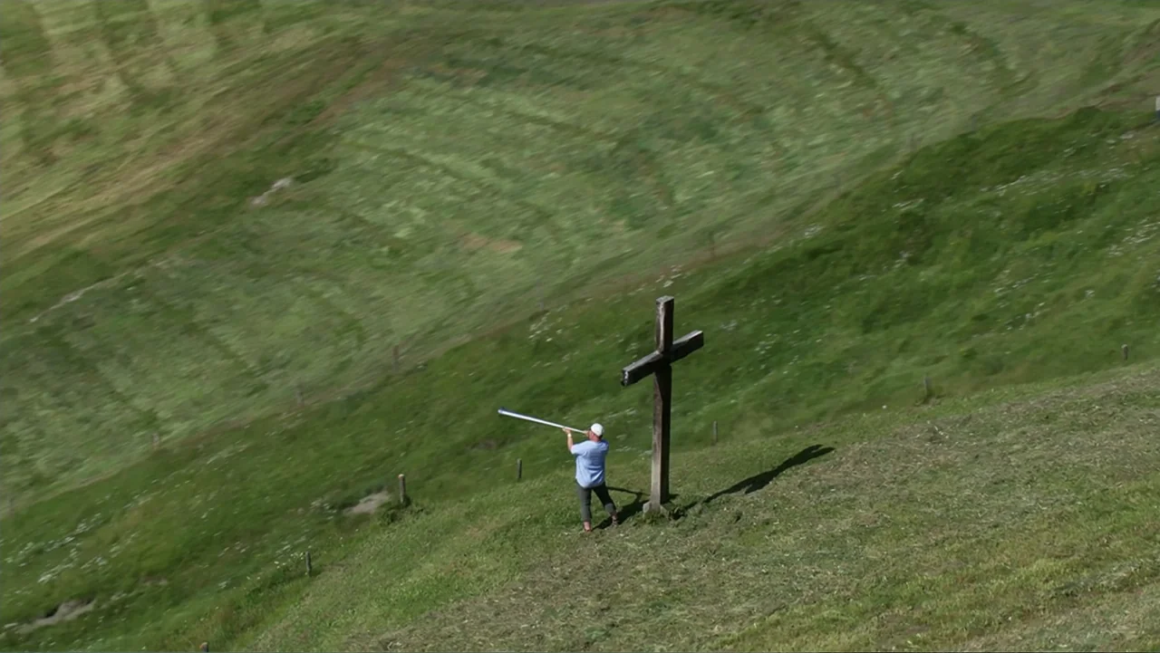 The Alps from Above: Symphony of Summits (2014) movie still: wide — A vast, rolling green hillside features a single large wooden cross near a path.…