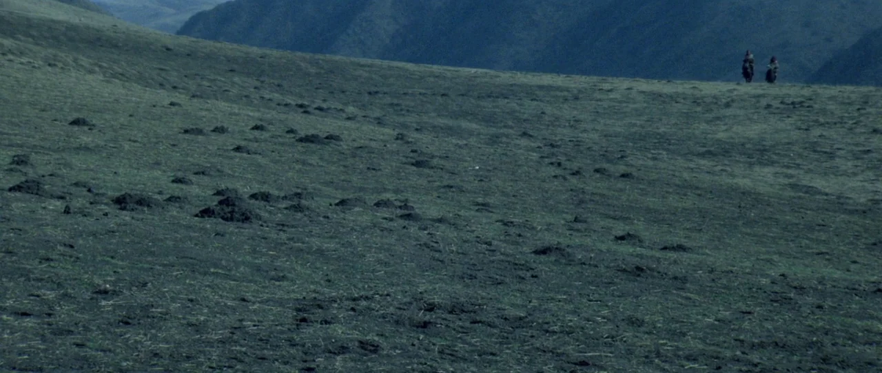 Horse Thief movie still: establishing — A vast, rolling grassland terrain stretches out under a mountain ridge in the distance. Two small figures on…