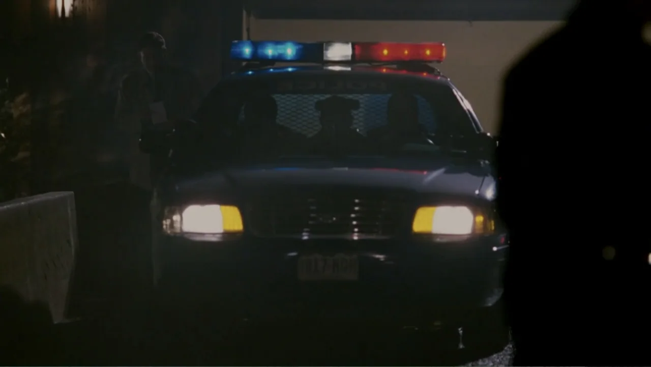 When a Stranger Calls (2006) movie still: medium — A police patrol car sits on a dark street at night with its lights flashing. An officer stands to…