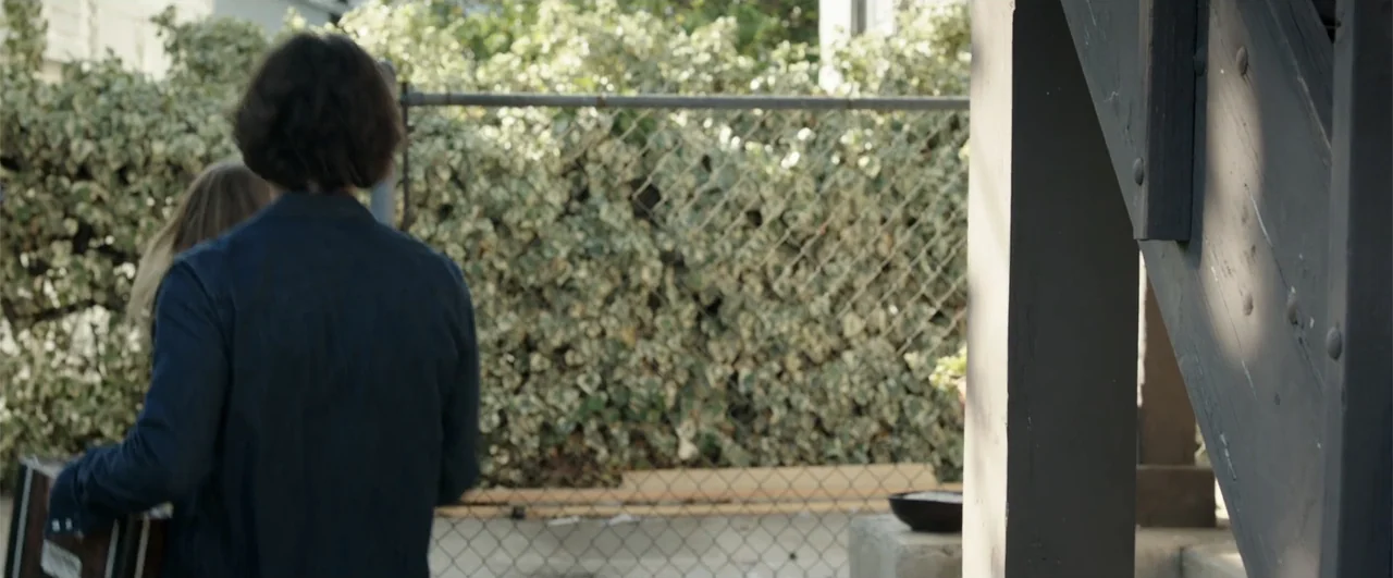 Lights Out (2016) movie still: medium — A chain-link fence covered in thick ivy borders an outdoor residential area. A man walks away from the…