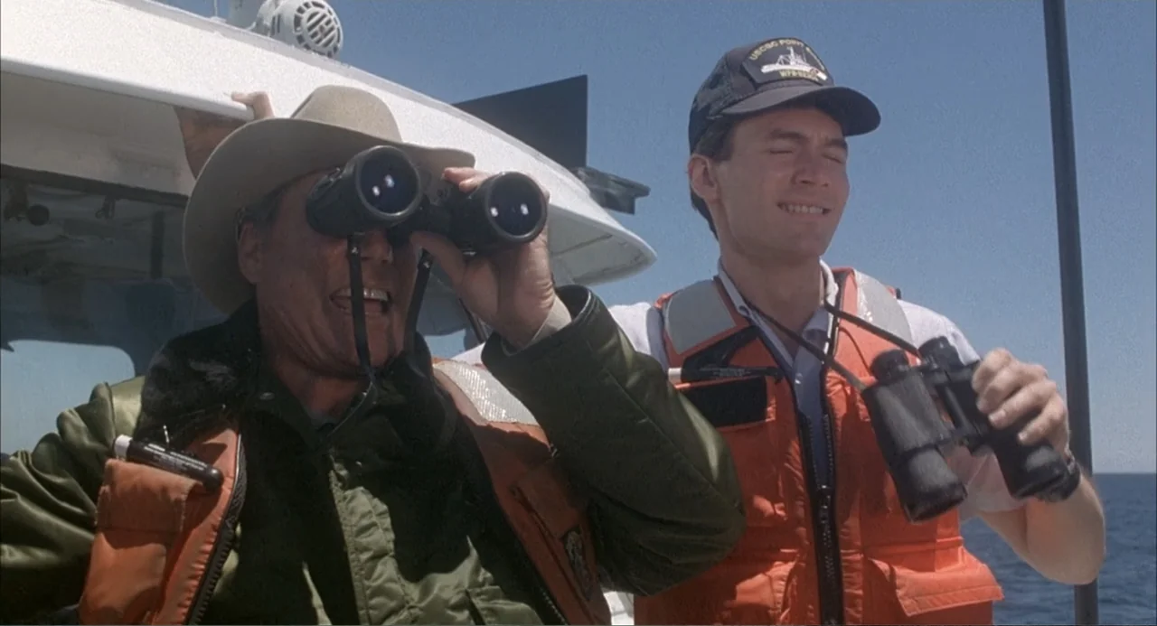 Overboard (1987) movie still: medium — Two men stand on the deck of a boat under a clear blue sky. Both men wear bright orange life vests and scan…