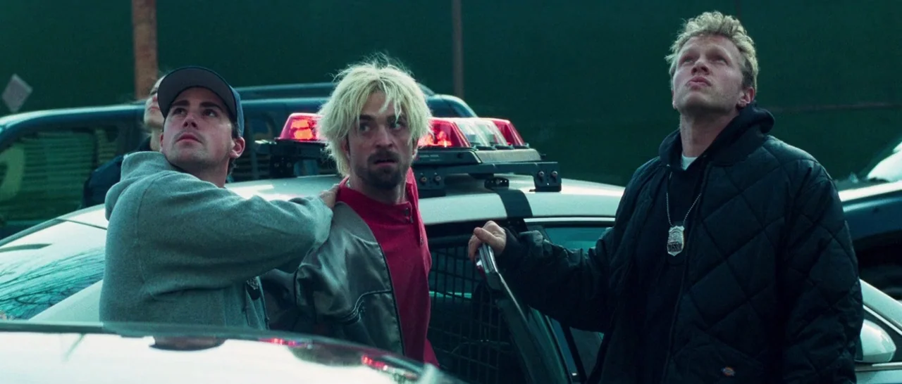 Good Time (2017) movie still: medium — A group of three men stand next to a police cruiser in a dark parking lot. The men look upward at an…