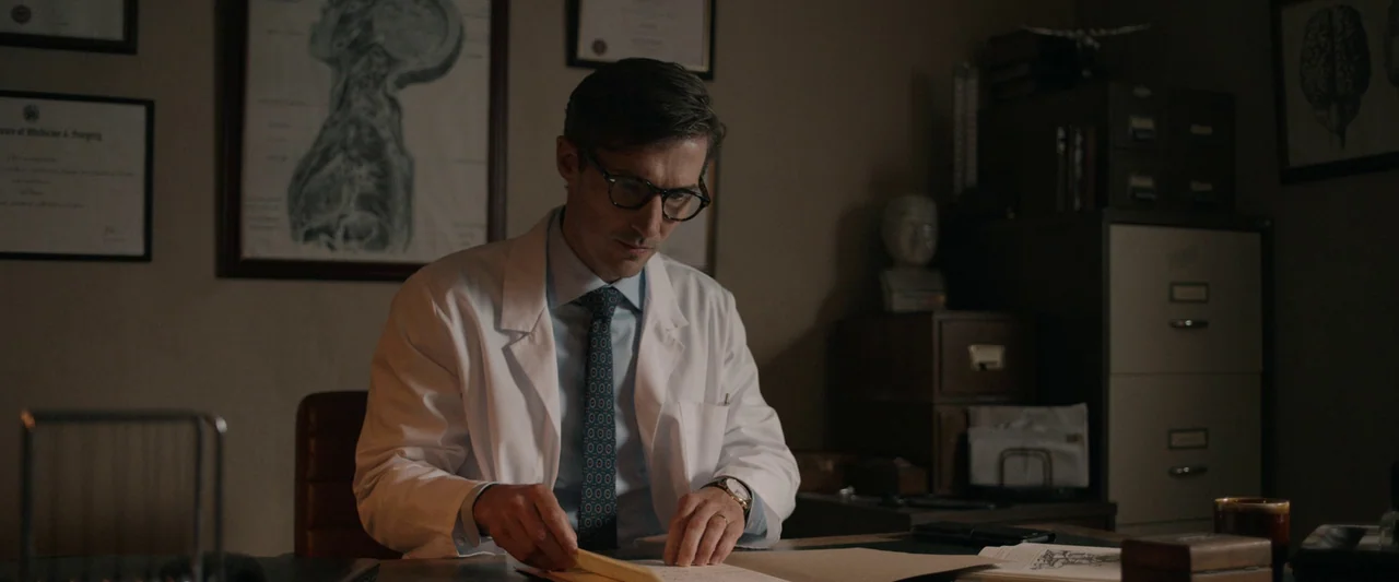 Oddity (2024) movie still: medium — A dimly lit room contains anatomical charts on the walls and metal filing cabinets. A man in a medical coat sits…