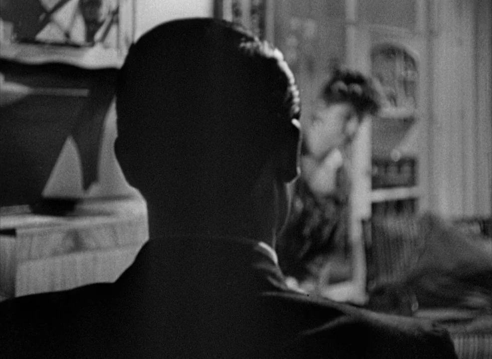 Notorious (1946) movie still: closeup — A dark indoor setting with a man in the foreground and a woman nearby. The man is sitting while the woman…