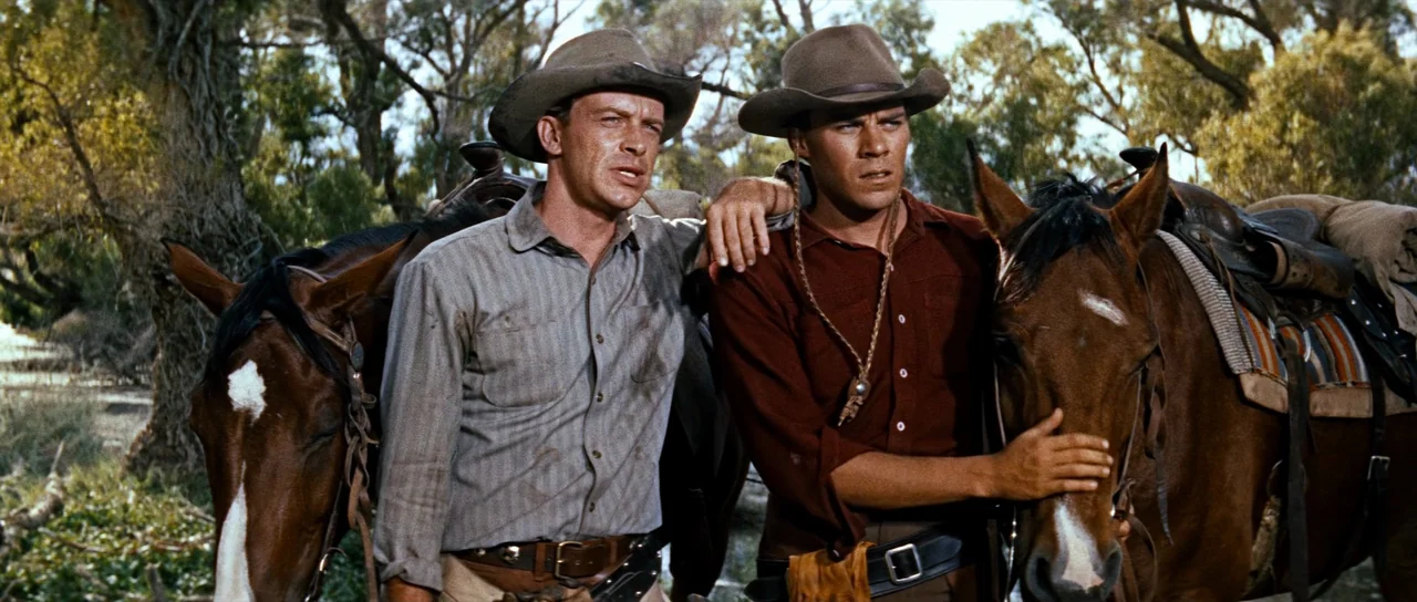 Comanche Station (1960) movie still: medium — Two men stand beside horses in a dense, sun-dappled grove of eucalyptus trees. The men are dressed in…