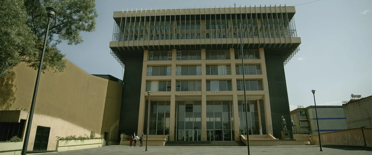Divine Intervention (2026) movie still: wide — A brutalist concrete office building with a cantilevered top section stands in daylight. Three small…
