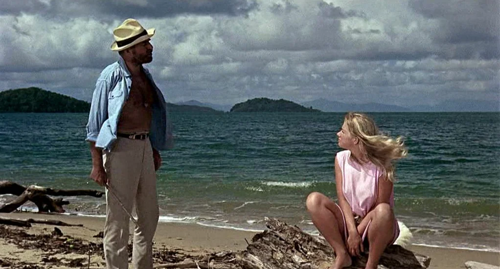 Age of Consent (1969) movie still: fullbody — A coastal shoreline with driftwood on the sand and dark forested hills across water. A man standing…