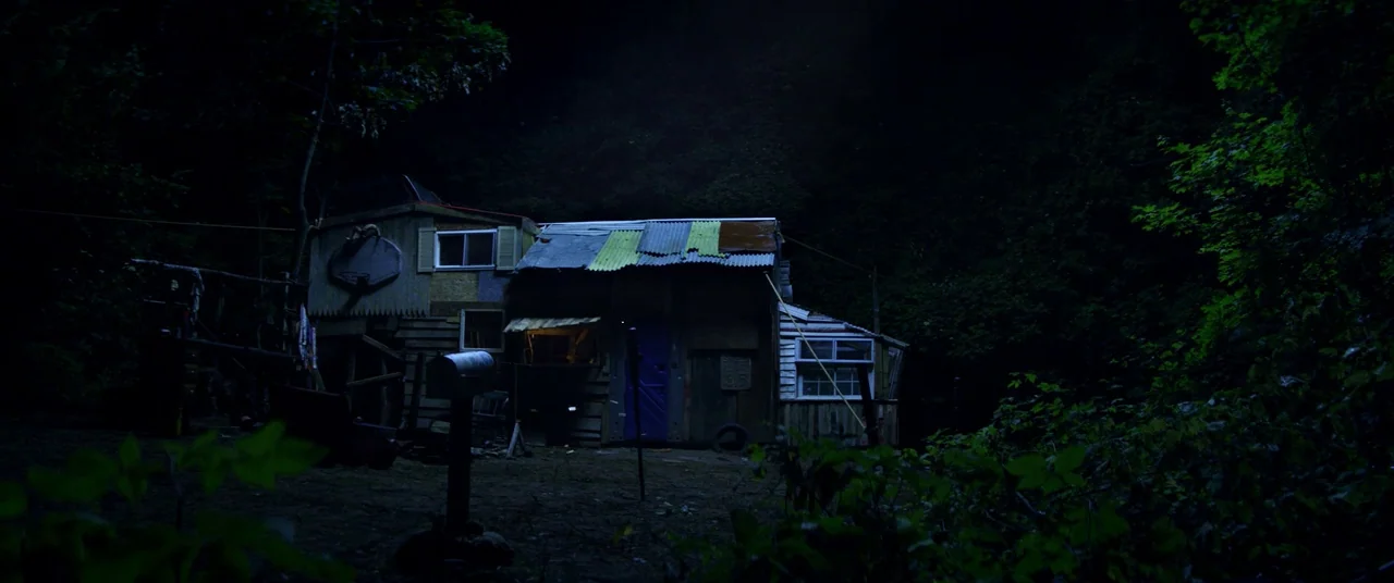 Kings Of Summer (2013) movie still: establishing — A makeshift wooden shack sits at the edge of a dark, dense forest at night. The structure is…