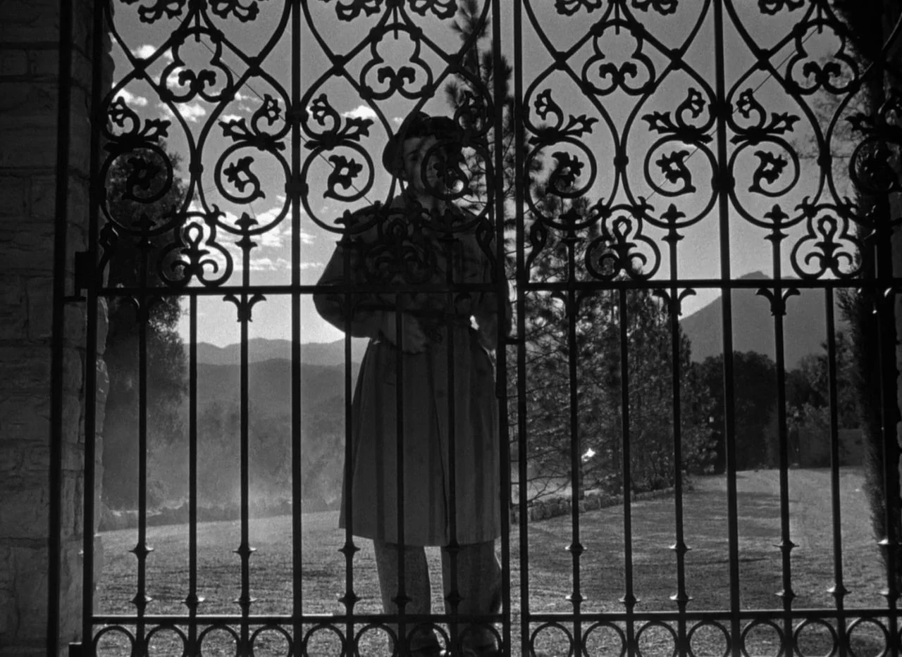 Out of the Past (1947) movie still: fullbody — A man stands behind an ornate black wrought iron gate leading to a field. Mountains are visible in…