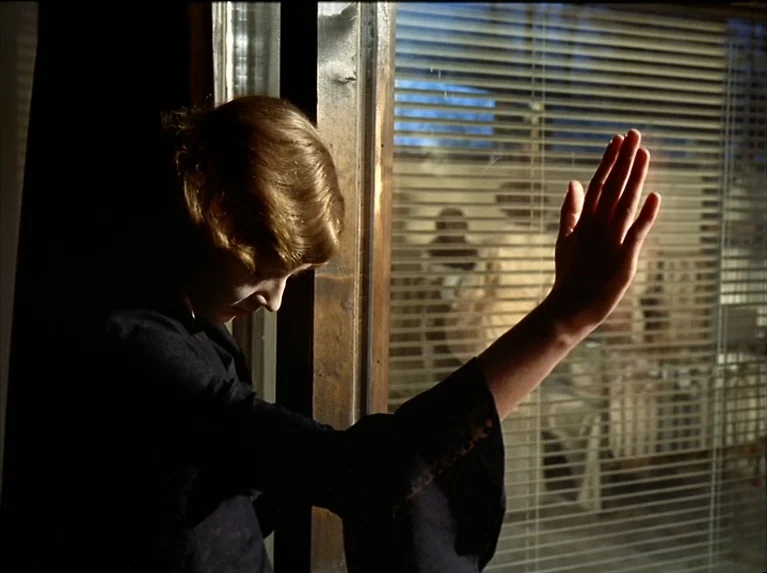 The Bitter Tears Of Petra Von Kant (1972) movie still: medium — A dimly lit interior room features a window with horizontal metal blinds. A young…