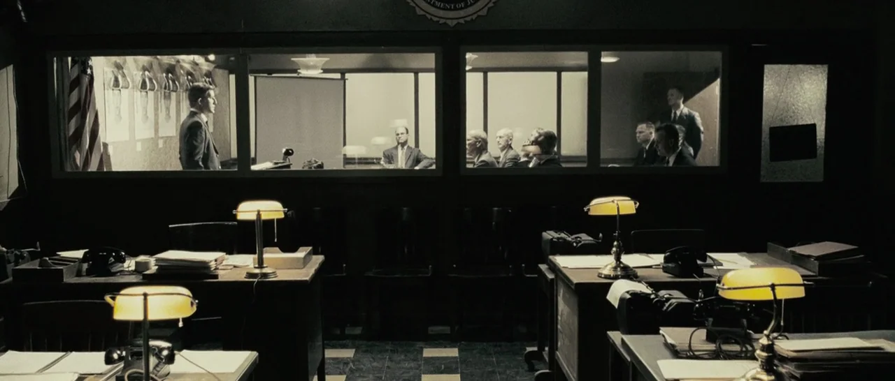 Slice (2018) movie still: wide — An observation room with dark wooden desks and glowing green banker lamps in foreground. A group of men stands and…