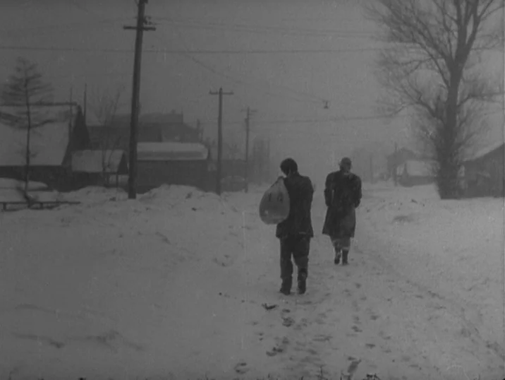 The Idiot (1951) movie still: wide — A snow-covered rural landscape with wooden houses and utility poles during a winter storm. Two figures in dark…