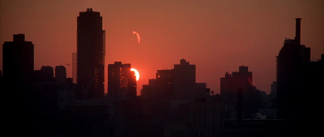 Die Hard with a Vengeance (1995) movie still: establishing — A dense urban skyline is silhouetted against a deep red sun setting behind buildings.…