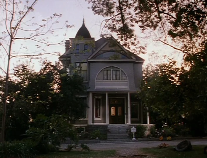 Head of the Family (1996) movie still: establishing — A two-story Victorian home with a turret stands behind leafy trees at dusk. A paved driveway…