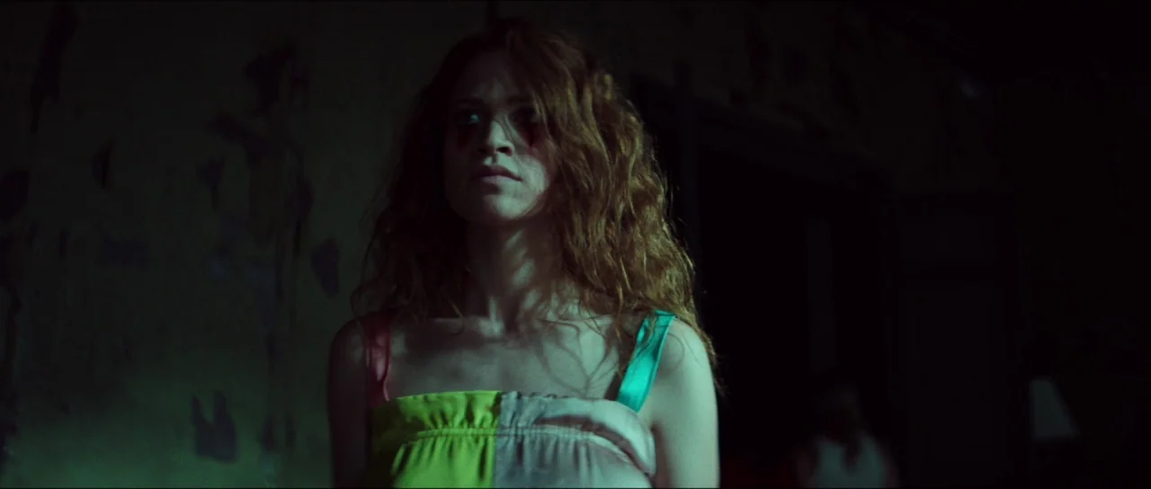 Braid (2018) movie still: medium — A dimly lit, decaying room features walls with peeling paint and shadows. A woman with long wavy hair stands in…