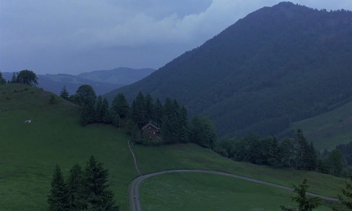 Phenomena (1985) movie still: establishing — A vast mountain landscape features a small house surrounded by dense coniferous trees. A winding gravel…