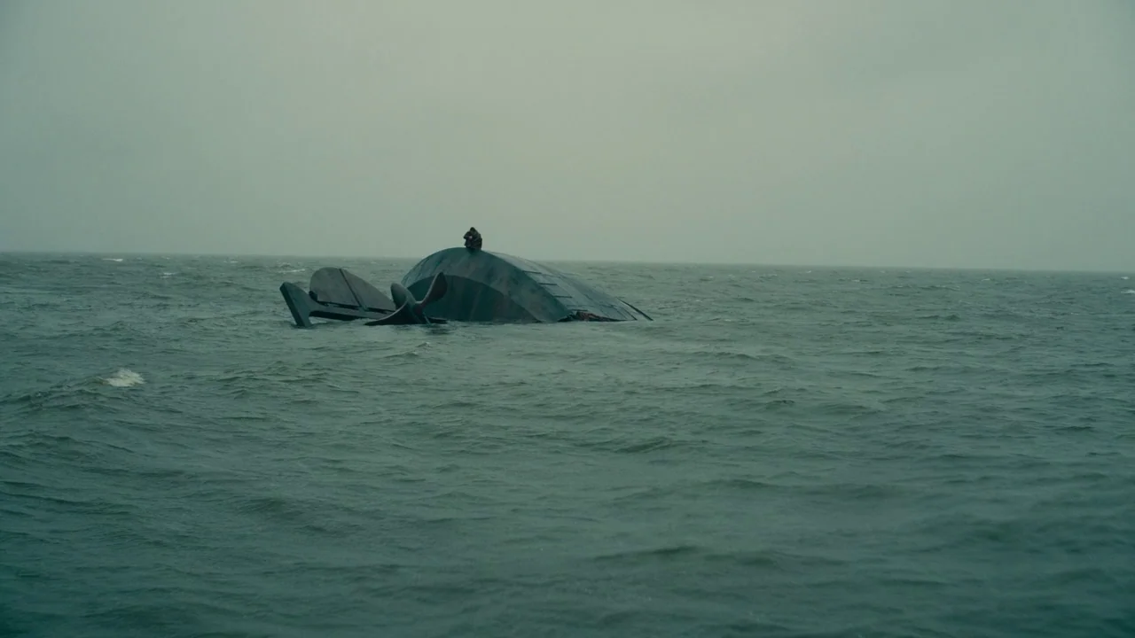 Dunkirk (2017) movie still: wide — A partially submerged, overturned boat hull drifts in the middle of a choppy ocean. A single small human figure…