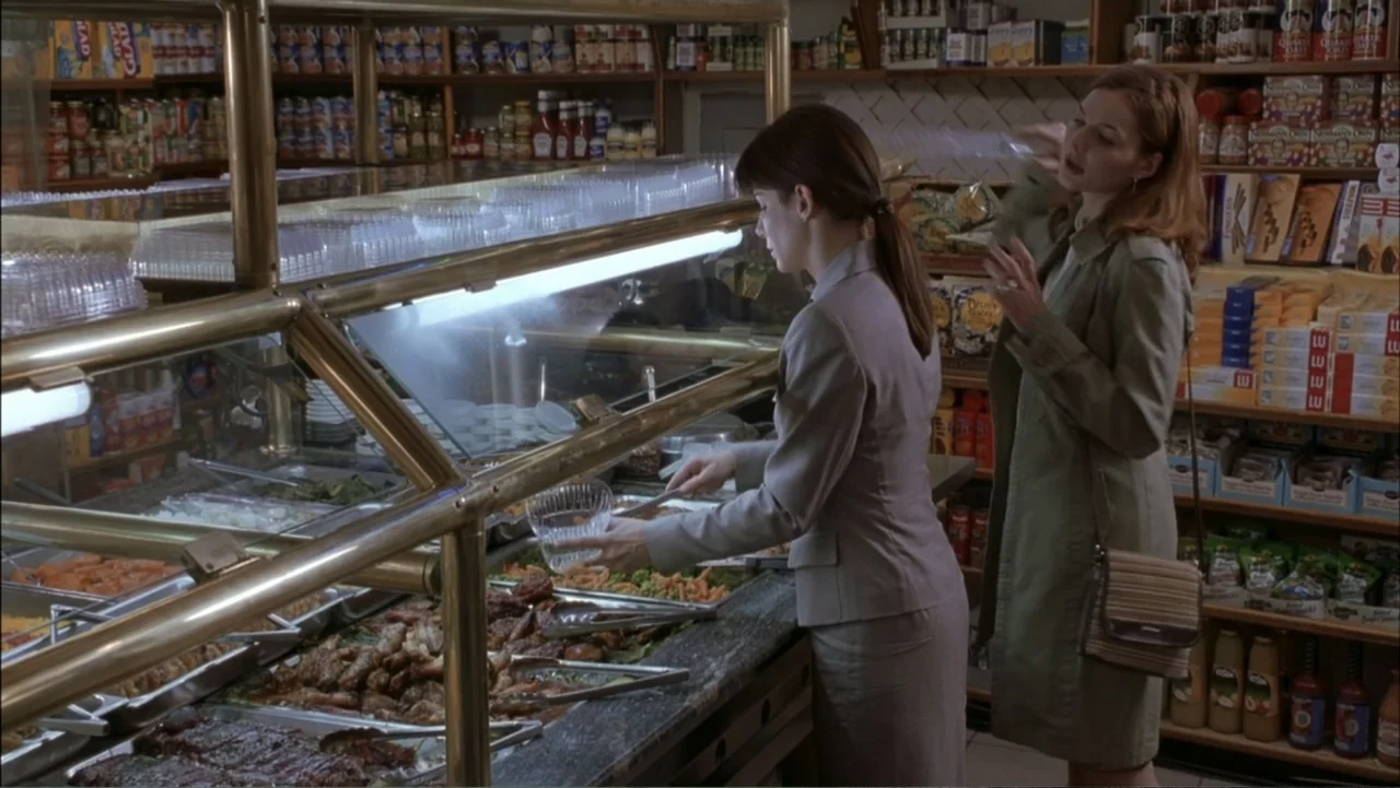 Two Weeks Notice (2002) movie still: medium — A food counter displays prepared meats and sides inside a grocery retail space. One woman in a grey…