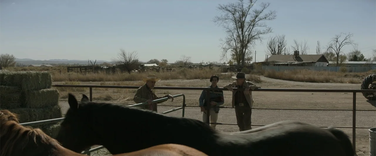 Cry Macho (2021) movie still: fullbody — A rural ranch corral with a metal fence and dirt ground under a clear sky. Three men stand together leaning…