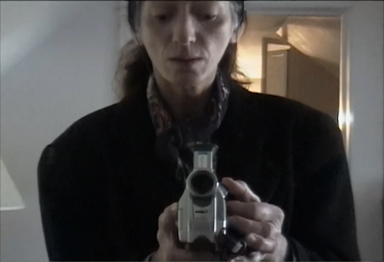 Plastic Disasters (2006) movie still: medium — An interior space contains a person seen through a reflection in a mirror. The woman holds a handheld…