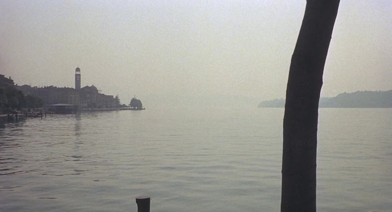 Salò, or the 120 Days of Sodom (1975) movie still: wide — A calm lake stretches toward a distant shoreline featuring a bell tower and buildings. A…