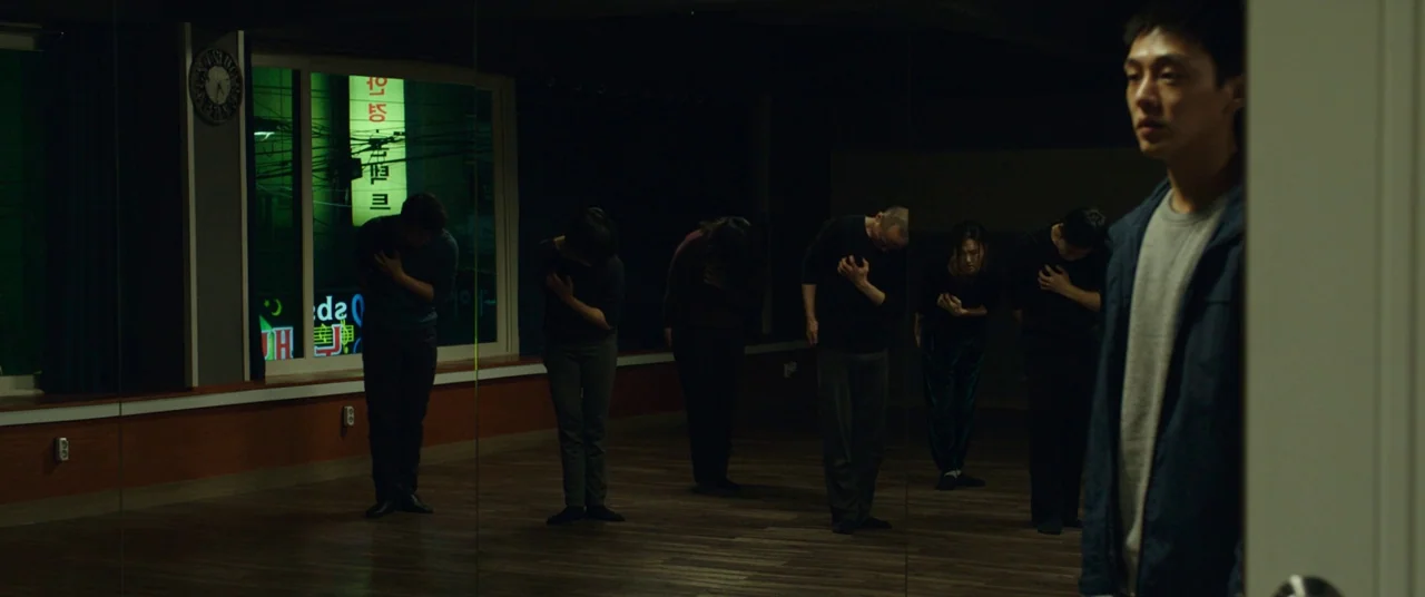 Burning (2018) movie still: wide — A dark dance studio with wooden floors and large mirrors covering the back wall. Five people stand in a line…