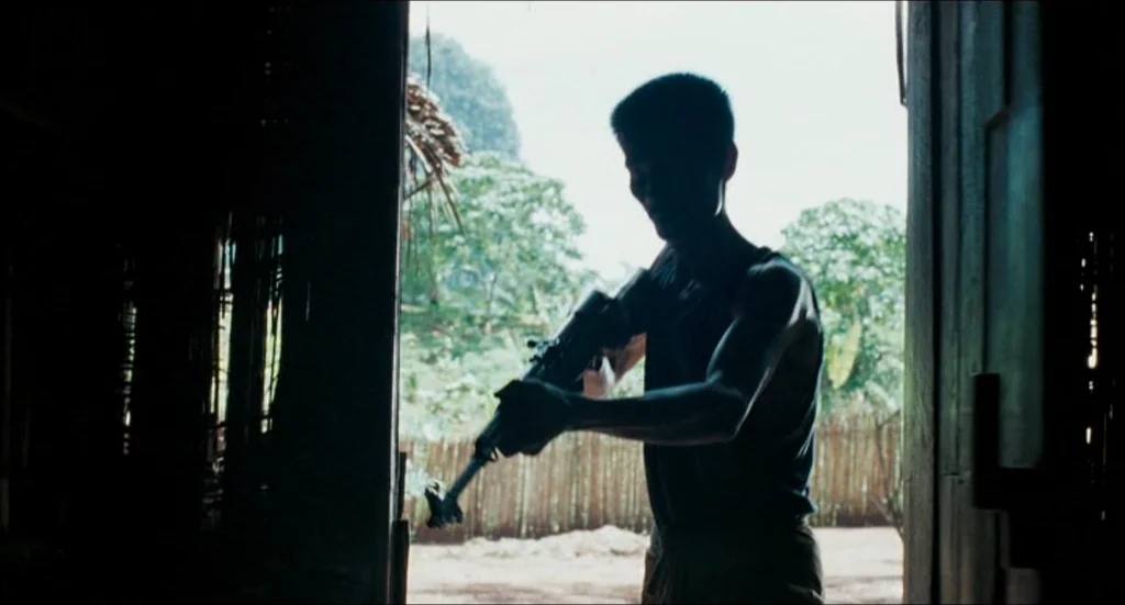 Rescue Dawn movie still: medium — A man stands in the dark doorway of a weathered wooden structure. He holds a rifle while looking toward the…