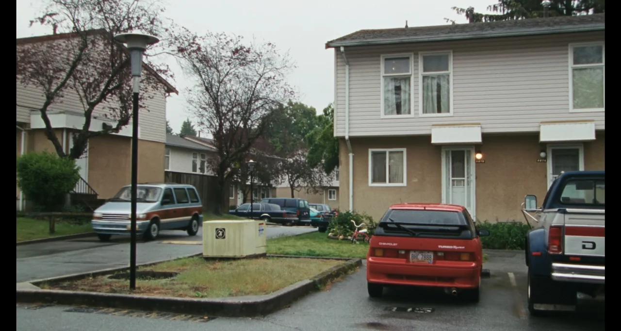 The Smashing Machine (2025) movie still: establishing — A quiet residential parking lot sits before two-story apartment buildings under a cloudy…