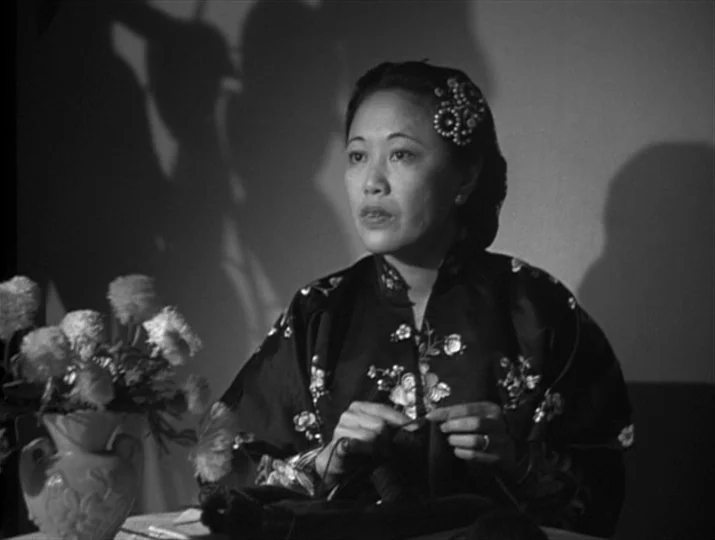 They Were Expendable (1945) movie still: medium — A woman sits indoors at night with a vase of flowers beside her. She is knitting while casting…