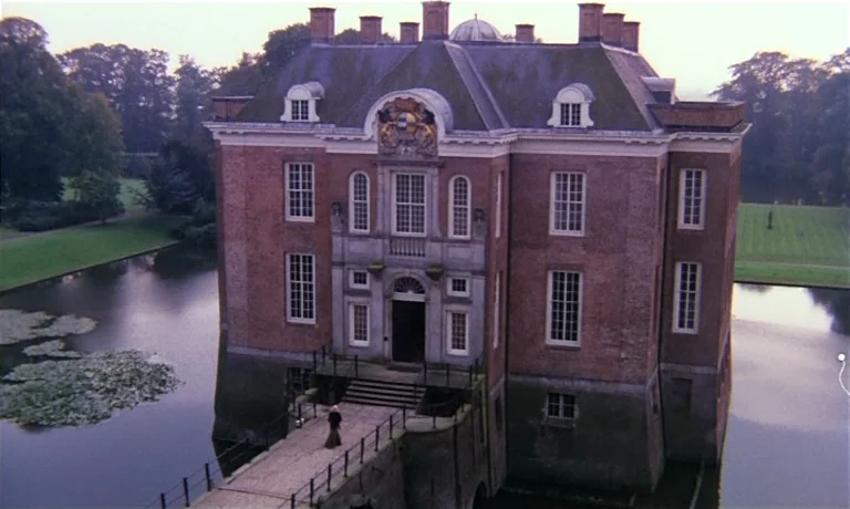 Katie Tippel (1975) movie still: establishing — A grand red-brick manor house sits surrounded by a wide dark moat. A person walks across the stone…
