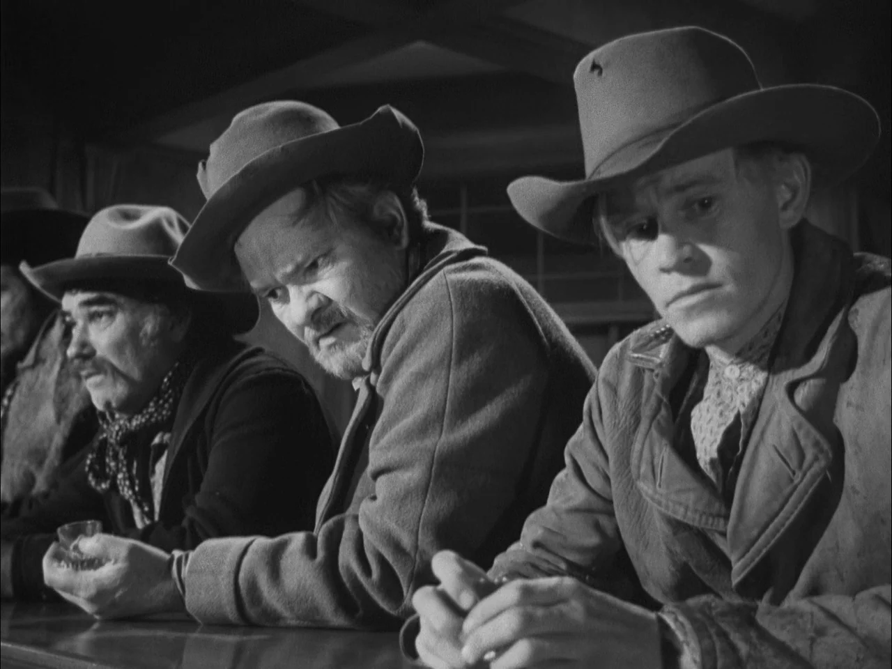The Ox-Bow Incident (1942) movie still: medium — Three men in western hats sit in a line at a dark wooden bar. They gaze downward as if distracted…