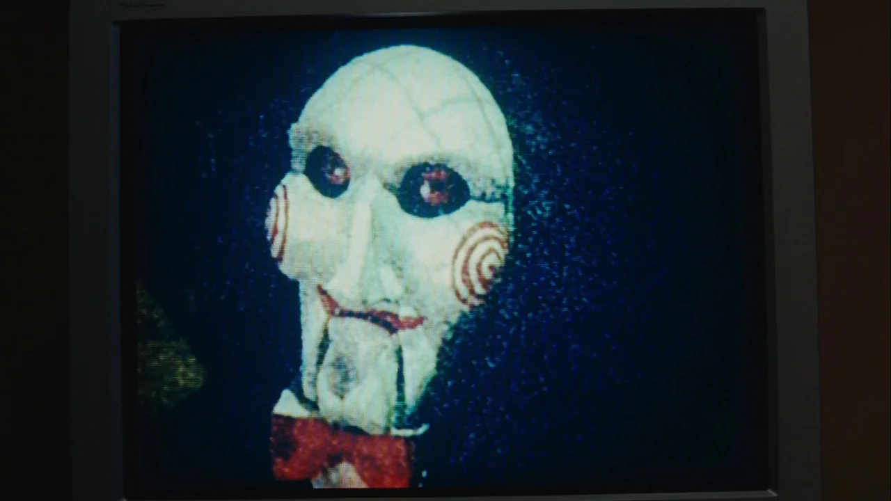Saw III (2006) movie still: closeup — A close-up view of a ventriloquist puppet displayed on a grainy television screen. The puppet has a white…