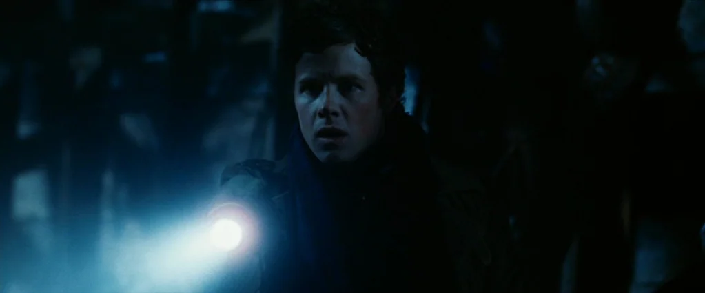 Wind Chill (2007) movie still: medium — A man stands in an underexposed environment, illuminated primarily by a bright handheld flashlight beam. The…