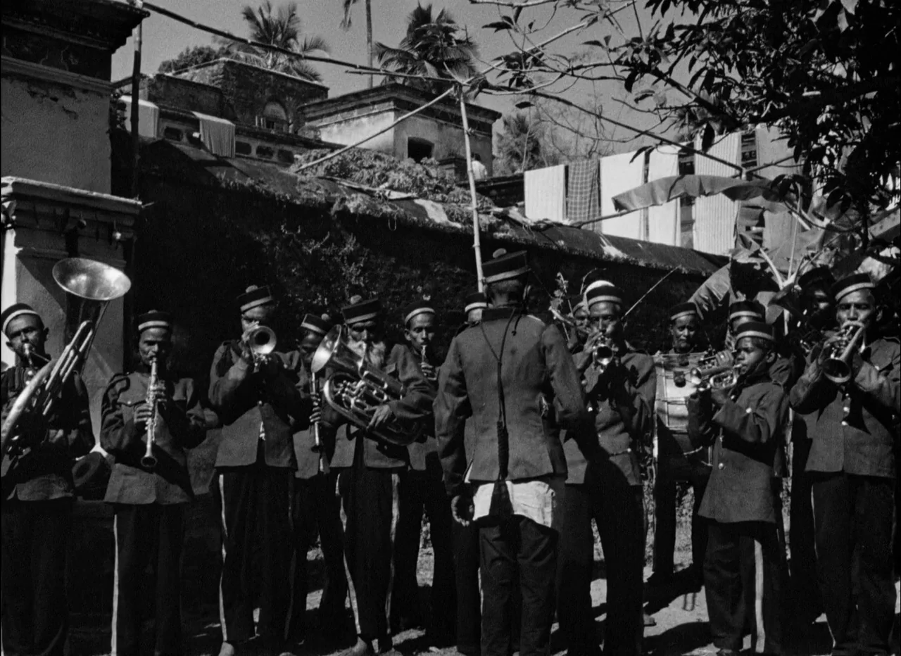 Pather Panchali (1955) movie still: fullbody — A brass band stands outdoors in a sunny, rural location beneath scattered tree branches. The men in…