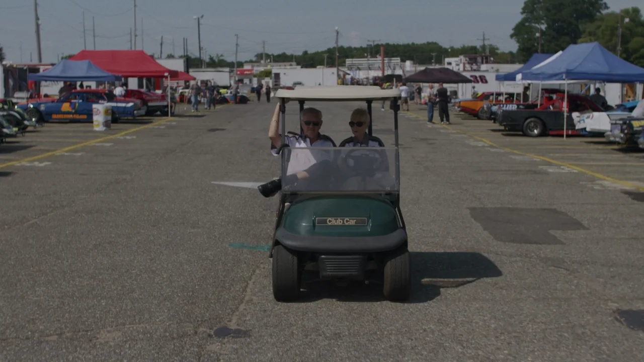 The Last Race (2022) movie still: medium — A paved racetrack paddock is lined with parked race cars and colorful canopy tents. An elderly couple…
