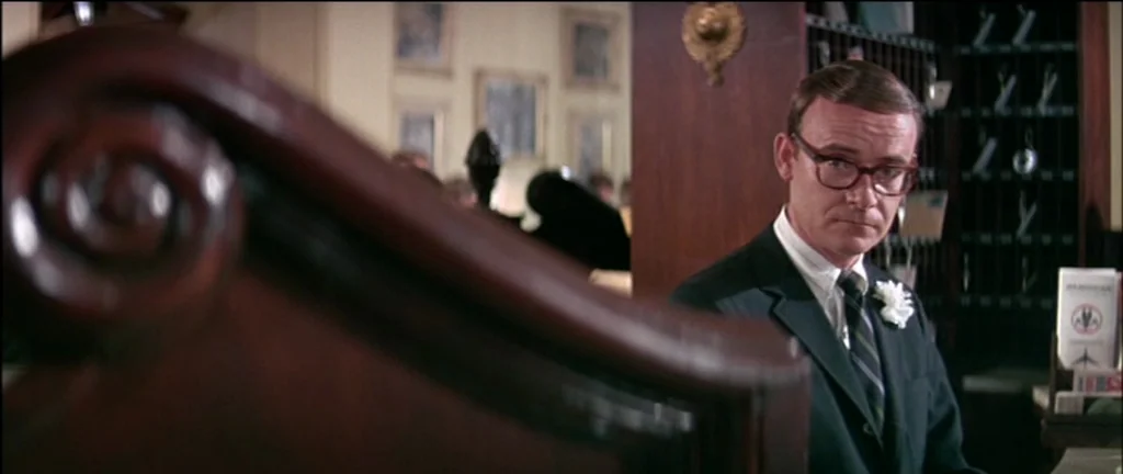 The Graduate (1967) movie still: medium — A wooden counter foreground obscures part of the view in a hotel lobby area. A man wearing a suit stands…