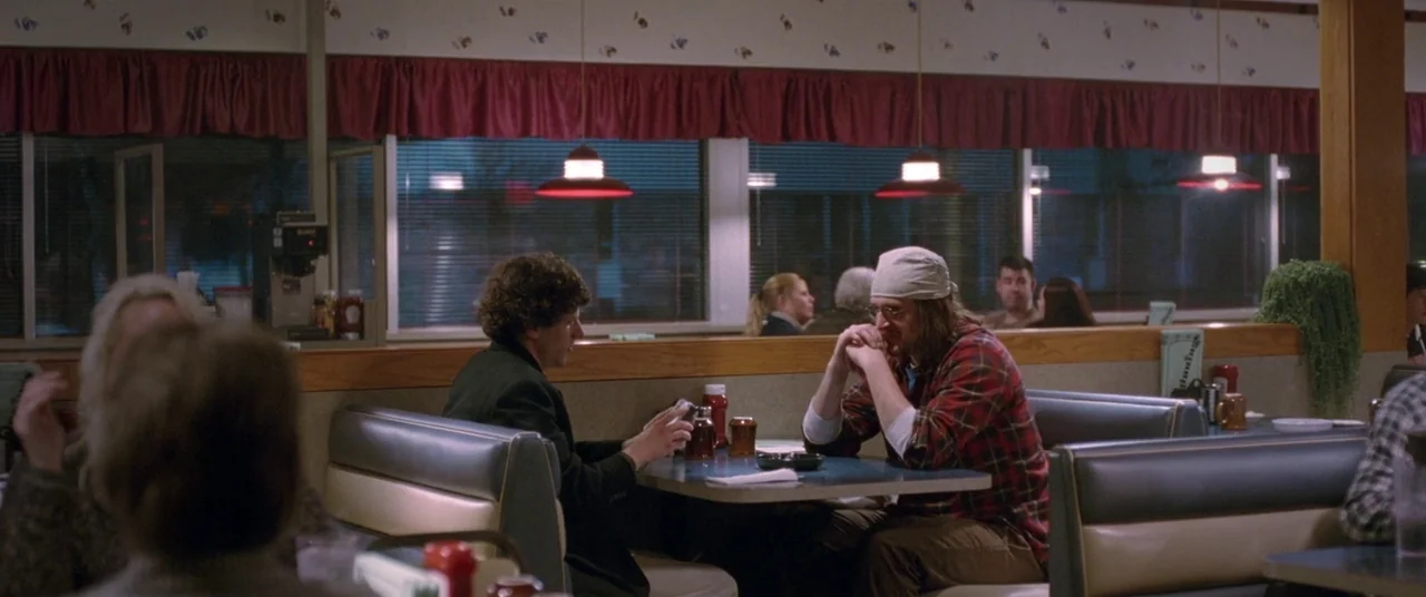 The End of the Tour (2015) movie still: medium — A diner booth with blue vinyl seats faces a long window overlooking a night exterior. Two men sit…