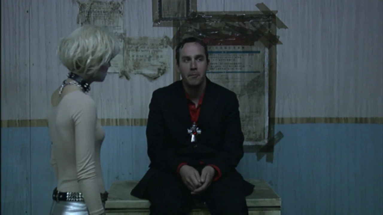 Murder Party movie still: medium — A workspace with plywood walls decorated with taped-up documents and blue lower paneling. A man in a suit sits on…