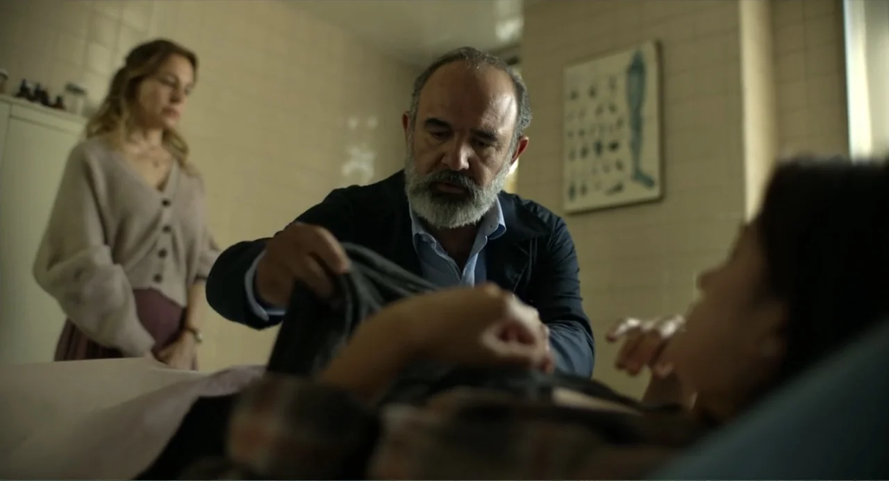 Heartbeat (2008) movie still: medium — A medical examination room features tiled walls and an anatomical chart on the wall. A man leans over a…