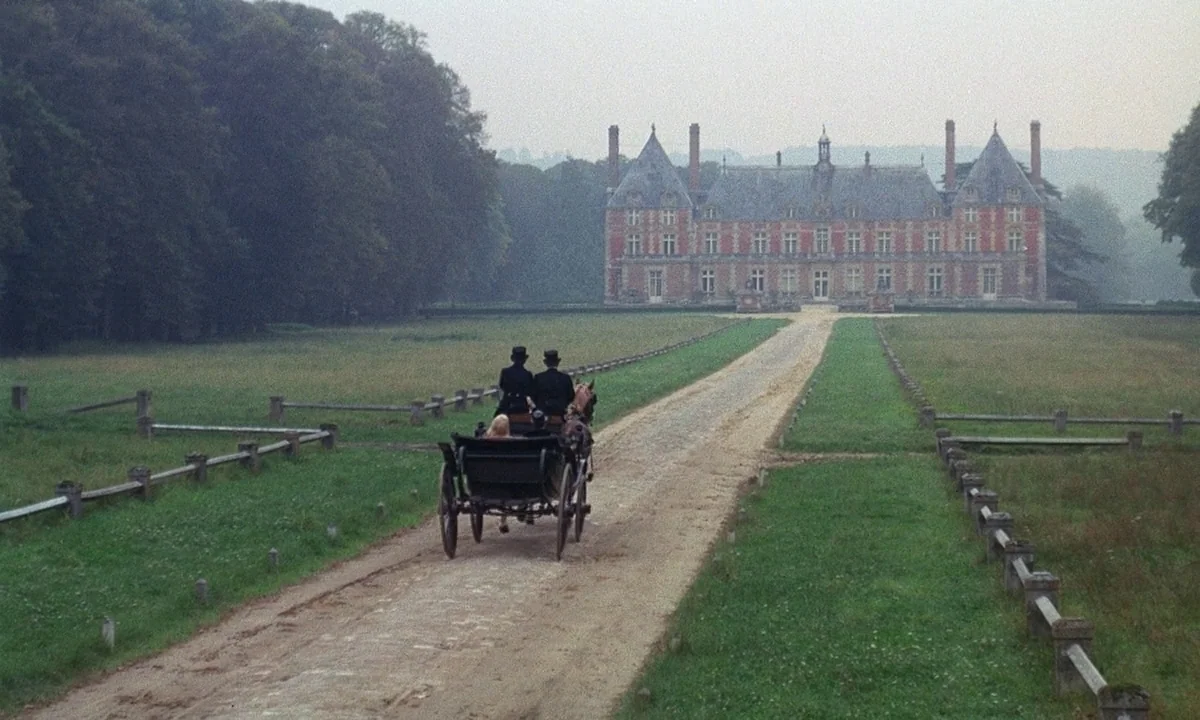 Belle De Jour (1967) movie still: wide — A grand, red-brick manor with symmetrical towers sits at the end of a dirt path. Two men in top hats ride…