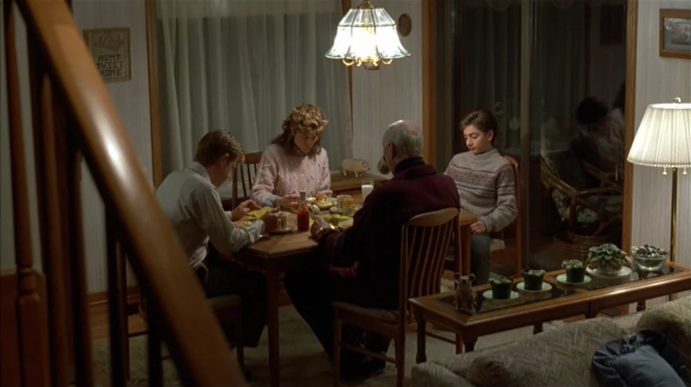 Fargo (1996) movie still: wide — A modest dining room features a wooden table and a decorative hanging light fixture. Four family members sit…