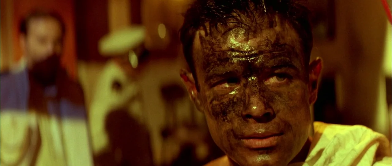 Querelle (1982) movie still: closeup — A dimly lit room bathed in yellow light appears cluttered and industrial. A man with a soot-covered face…